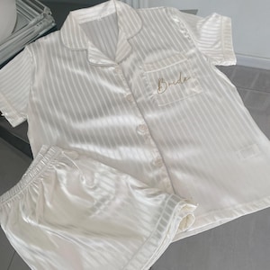 May include: White striped satin pyjama set with short sleeves and shorts. The top has a button-up front and a pocket on the left chest that says "Bride" in gold embroidery.