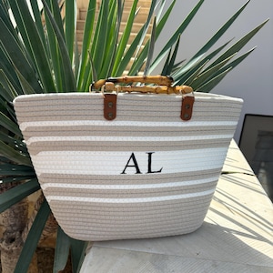 May include: A beige and white striped tote bag with a bamboo handle and leather accents. The bag has the initials "AL" embroidered on the front.