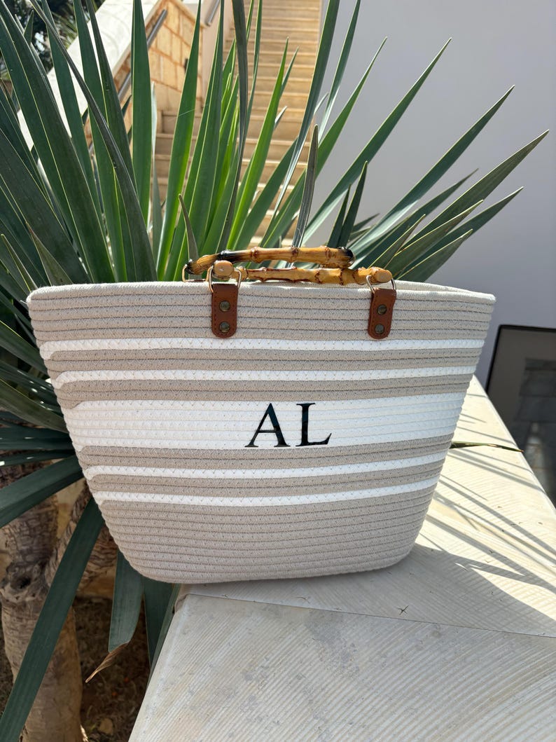 May include: A beige and white striped tote bag with a bamboo handle and leather accents. The bag has the initials "AL" embroidered on the front.
