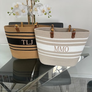 May include: Two striped tote bags with natural rope handles and leather accents. The bag on the left has black and tan stripes with the initials "TLJ" embroidered on the front. The bag on the right has white and tan stripes with the initials "MMO" embroidered on the front.