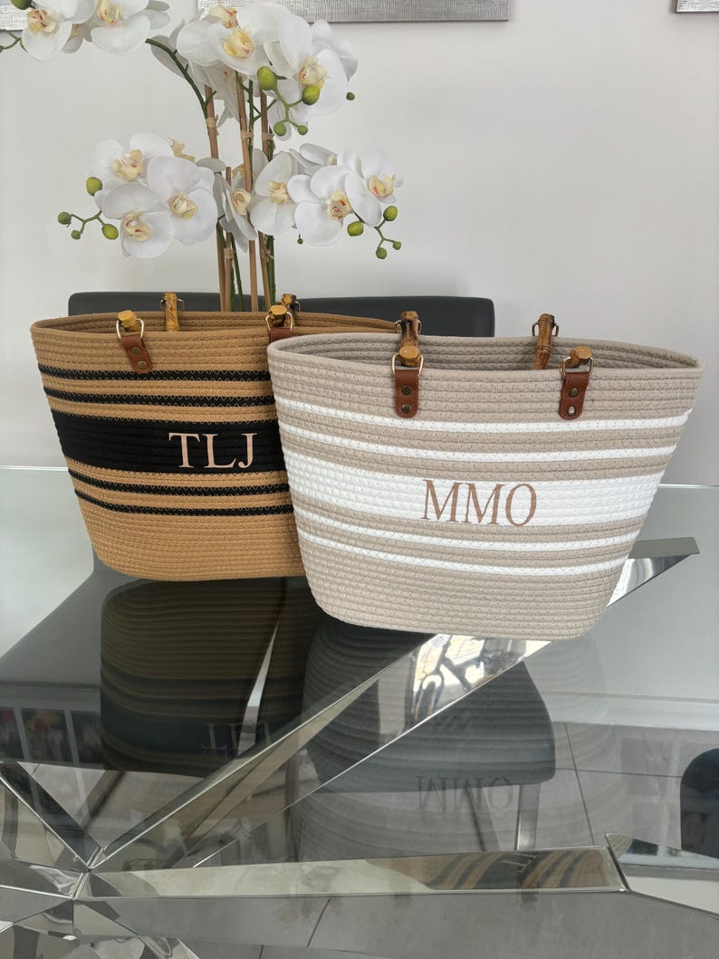 May include: Two striped tote bags with natural rope handles and leather accents. The bag on the left has black and tan stripes with the initials "TLJ" embroidered on the front. The bag on the right has white and tan stripes with the initials "MMO" embroidered on the front.