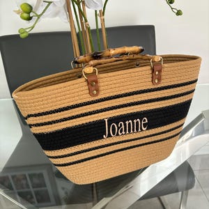 May include: A tan and black striped tote bag with bamboo handles. The bag has the name "Joanne" embroidered on the front.