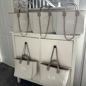 May include: Five beige canvas tote bags with chain straps and personalized initials. The initials are "EJM", "CL", "MML", "TFC", and "L.H."