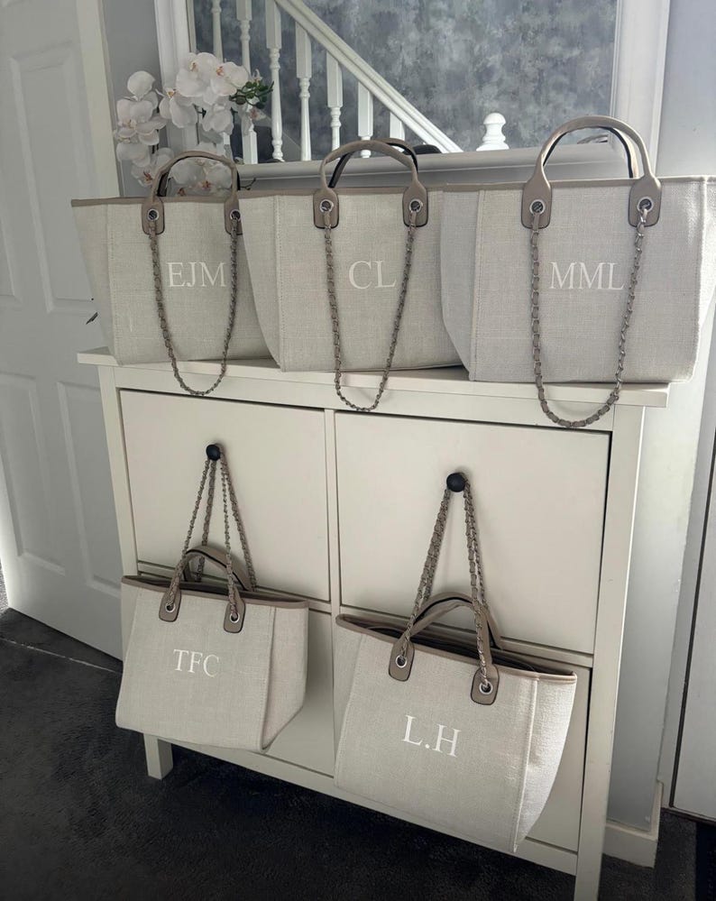 May include: Five beige canvas tote bags with chain straps and personalized initials. The initials are "EJM", "CL", "MML", "TFC", and "L.H."