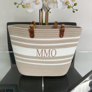 May include: A beige and white striped tote bag with brown leather handles and a wooden top. The bag has the initials "MMO" embroidered on the front.