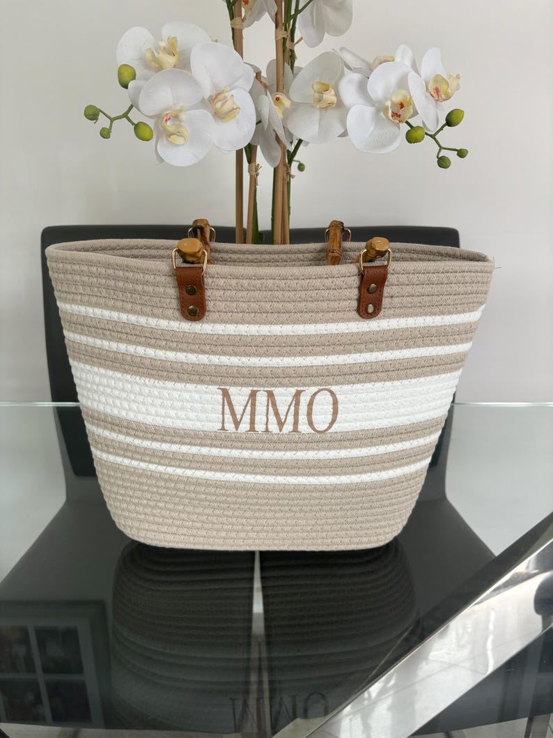 May include: A beige and white striped tote bag with brown leather handles and a wooden top. The bag has the initials "MMO" embroidered on the front.