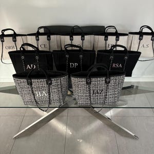 May include: A group of nine black and white tote bags with personalized initials on a glass table. The bags are made of canvas and have a chain strap. The initials are in a gold color.