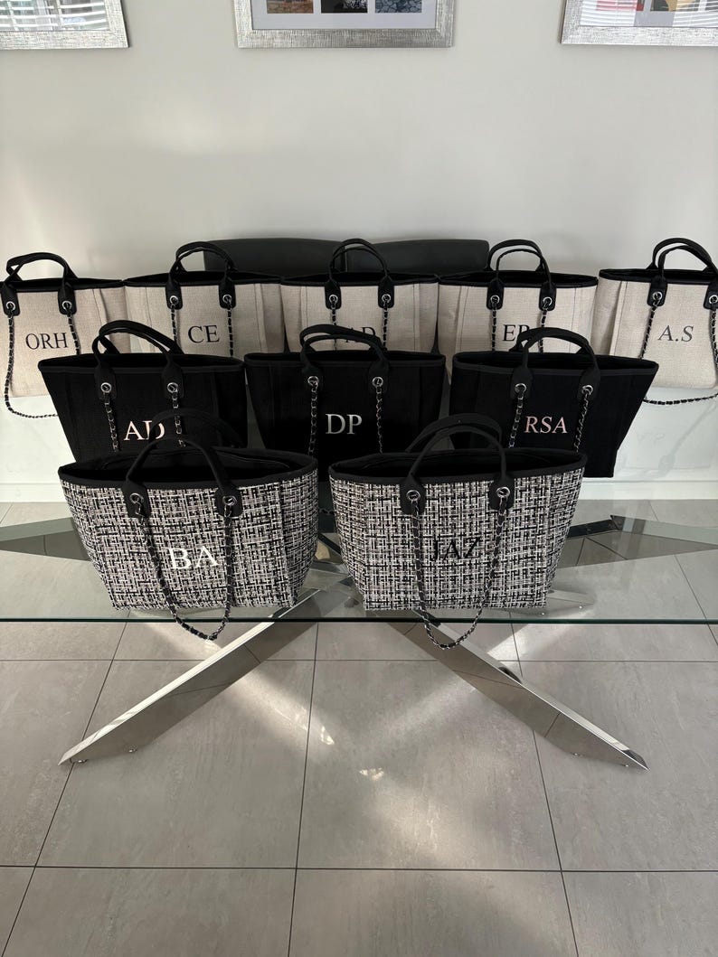 May include: A group of nine black and white tote bags with personalized initials on a glass table. The bags are made of canvas and have a chain strap. The initials are in a gold color.