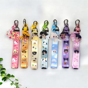 May include: A collection of seven colorful lanyards with various cartoon character charms. Each lanyard features a different design, including characters like Hello Kitty, foxes, and anime-style figures. The lanyards have metal clasps and are displayed against a white background.