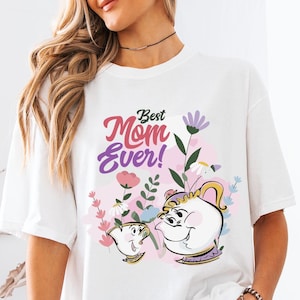 Könnte beinhalten: Weißes T-Shirt mit dem Text "Best Mom Ever!" in Rosa und Lila, mit einer Cartoon-Illustration einer Teekanne und einer Teetasse mit floralen Akzenten. Das Shirt hat kurze Ärmel und eine lockere Passform.