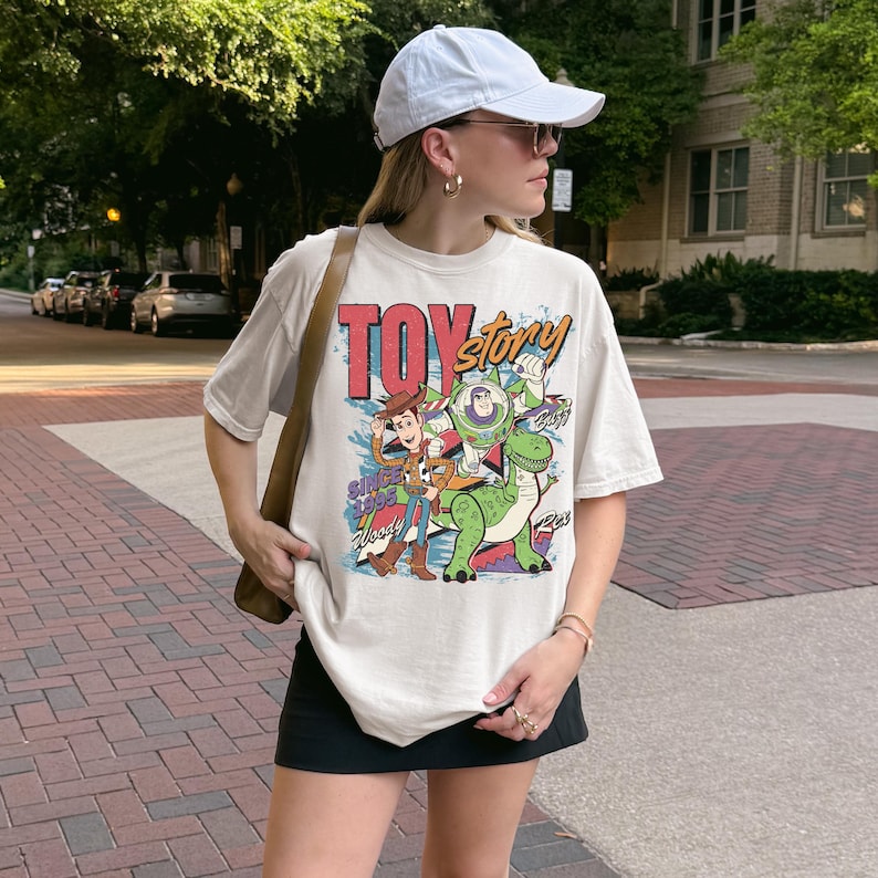 A woman wearing a white cap and a white t-shirt with a colorful graphic of characters from the Toy Story franchise stands on a brick sidewalk.