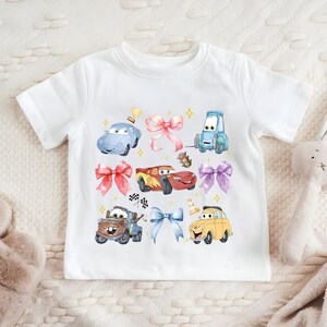 A white t-shirt with a colorful graphic of cartoon cars and a stuffed rabbit on it, along with various other cartoon-style objects.