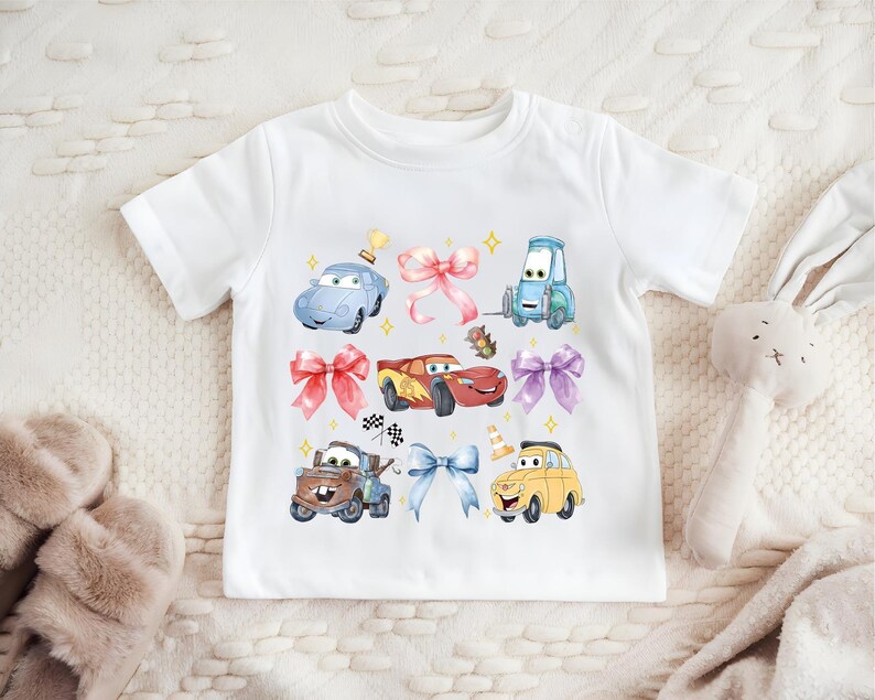 A white t-shirt with a colorful graphic of cartoon cars and a stuffed rabbit on it, along with various other cartoon-style objects.