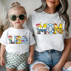 May include: White t-shirts with colorful graphic designs. One shirt says "LISA" with cartoon characters, and the other says "MOM" with cartoon characters. The shirts are worn by a child and an adult.