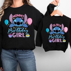 A woman wearing a black sweatshirt with a design featuring a cartoon character and text that says "Mommy Birthday Girl". The woman is holding the sweatshirt up to the camera.