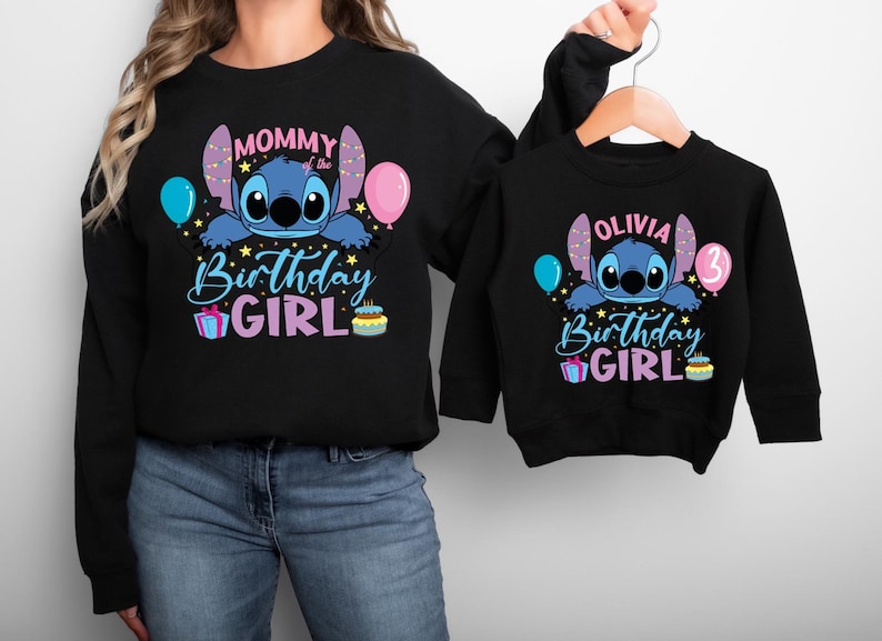 A woman wearing a black sweatshirt with a design featuring a cartoon character and text that says "Mommy Birthday Girl". The woman is holding the sweatshirt up to the camera.