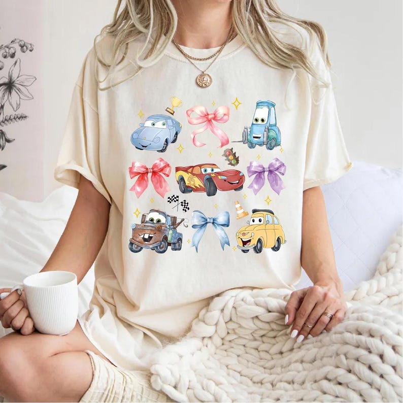 A woman wearing a white t-shirt with a graphic of cartoon cars and various other objects, including a cup and a blanket.