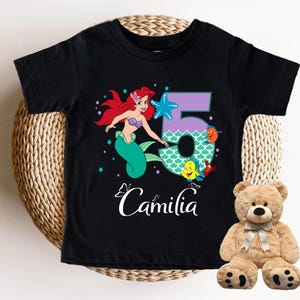 A black t-shirt with a cartoon mermaid character and the number 5, along with the text "Camila" printed on it. Next to the t-shirt is a stuffed teddy bear.