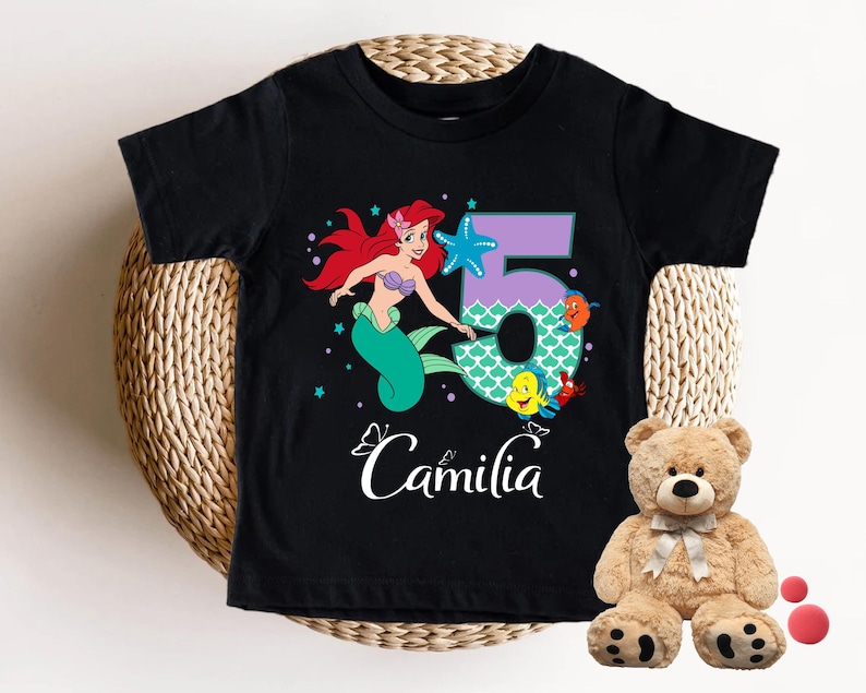 A black t-shirt with a cartoon mermaid character and the number 5, along with the text "Camila" printed on it. Next to the t-shirt is a stuffed teddy bear.