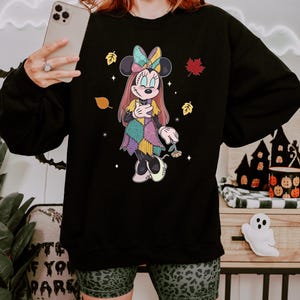 A woman wearing a black sweatshirt with a Minnie Mouse design is holding a smartphone and smiling.