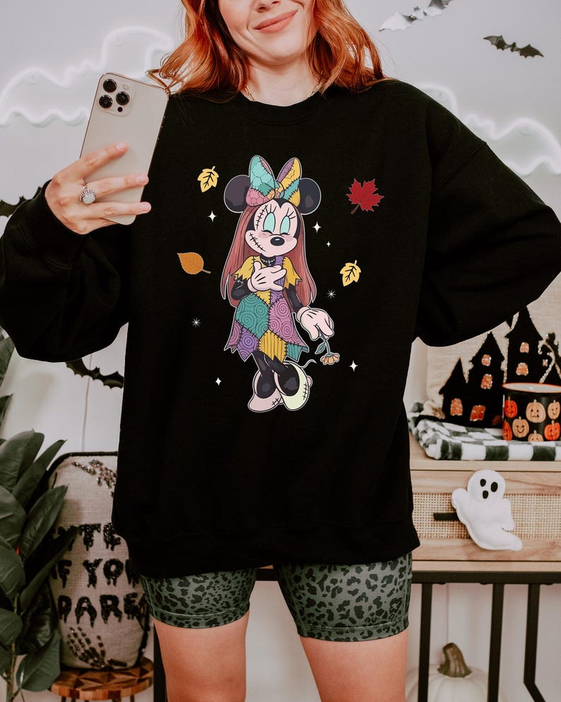 A woman wearing a black sweatshirt with a Minnie Mouse design is holding a smartphone and smiling.