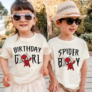 Two children wearing white t-shirts with the text "BIRTHDAY GIRL" and "SPIDER BOY" printed on them, smiling and posing together outdoors.