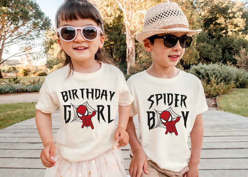 Two children wearing white t-shirts with the text "BIRTHDAY GIRL" and "SPIDER BOY" printed on them, smiling and posing together outdoors.