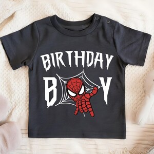 A black t-shirt with a Spiderman graphic and the text "BIRTHDAY B" printed on it, along with a pair of sandals and a stuffed rabbit.