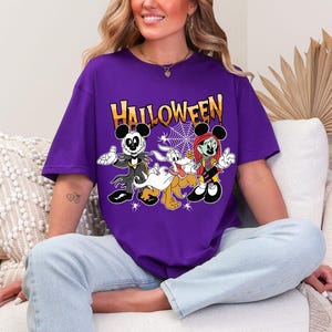 A woman wearing a purple t-shirt with a Halloween-themed graphic of Mickey Mouse and other characters is sitting on a couch.