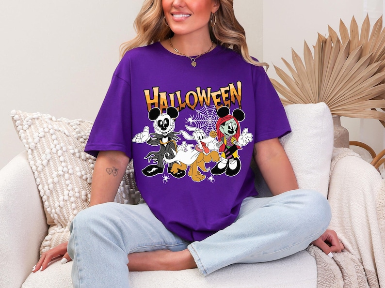 A woman wearing a purple t-shirt with a Halloween-themed graphic of Mickey Mouse and other characters is sitting on a couch.