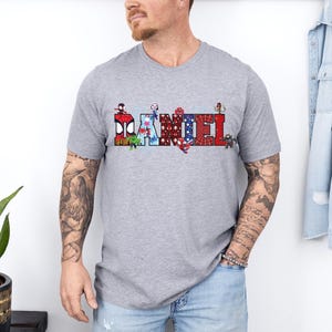 A man wearing a gray t-shirt with a graphic design of various characters on it, standing in front of a white wall.