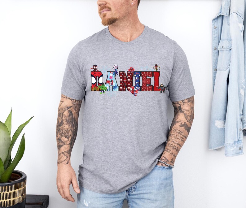 A man wearing a gray t-shirt with a graphic design of various characters on it, standing in front of a white wall.