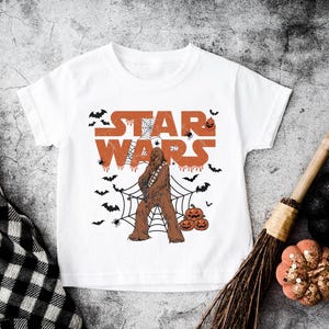 A white t-shirt with a Star Wars character and Halloween-themed decorations, including bats, pumpkins, and a broom, is displayed on a textured surface.