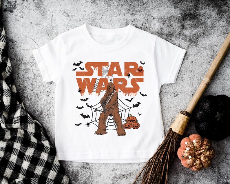 A white t-shirt with a Star Wars character and Halloween-themed decorations, including bats, pumpkins, and a broom, is displayed on a textured surface.