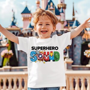 A young boy wearing a white t-shirt with the text "SUPERHERO SQUAD" printed on it, smiling and having his arms outstretched in front of a colorful, castle-like structure.