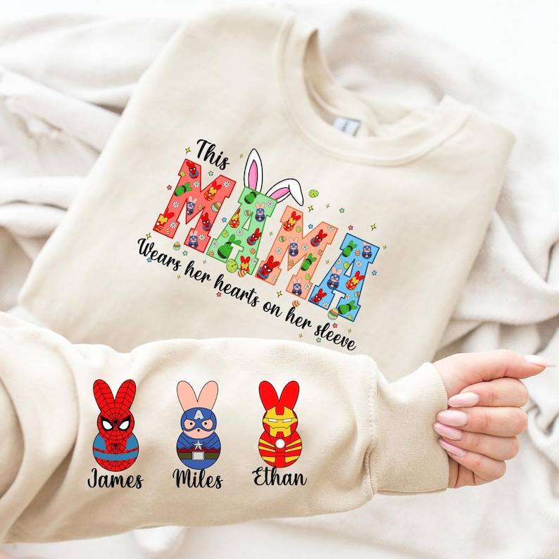 A person is holding a white t-shirt with a colorful design featuring the letters "MAMMA" and four cartoon rabbit characters.
