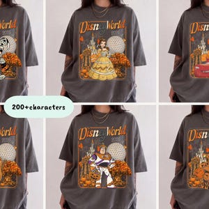 May include: A charcoal grey short-sleeve t-shirt featuring a graphic print with the text Disney World 2026. The design includes various Disney characters, the Epcot sphere, and autumn-themed elements like pumpkins and orange trees in a square frame.