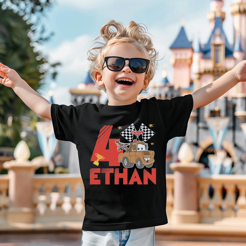A young boy with curly hair and sunglasses is smiling and raising his arms, wearing a black t-shirt with a graphic of a race car and the number 4.