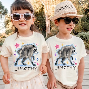Pode incluir: Duas t-shirts de manga curta na cor creme com uma ilustração gráfica de um guaxinim e o nome JIMOTHY impresso a preto por baixo do animal. O design inclui detalhes de estrelas coloridas em rosa, azul e amarelo à volta do guaxinim.