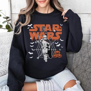 A woman wearing a black sweatshirt with a Star Wars-themed graphic design, including a stormtrooper, spider web, and Halloween pumpkins.