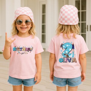 A young girl wearing a pink t-shirt with a birthday message and a pink and white checkered hat, smiling and making a hand gesture.