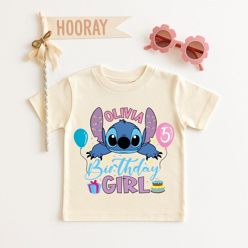 A white t-shirt with a cartoon character and text, along with a banner that says "Hooray" and pink sunglasses, all arranged on a white background.