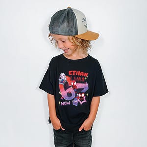A young boy wearing a black t-shirt with a graphic design and a baseball cap, smiling and looking down.