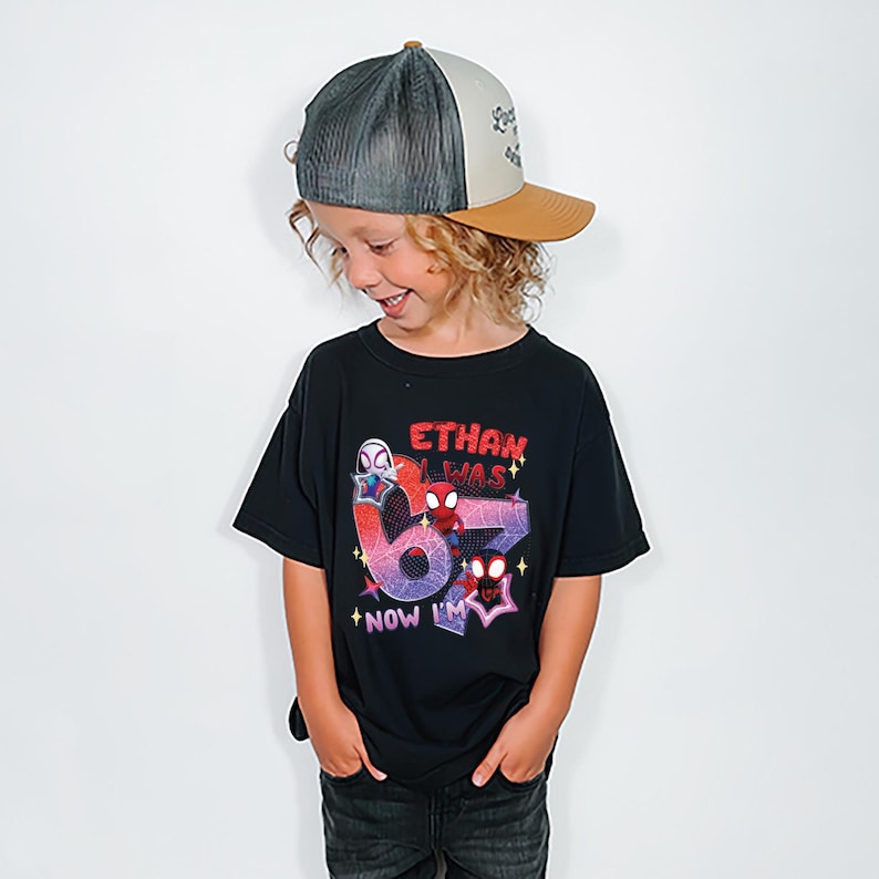 A young boy wearing a black t-shirt with a graphic design and a baseball cap, smiling and looking down.