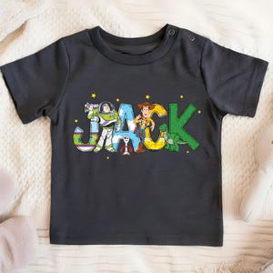May include: Dark gray short-sleeved t-shirt featuring the name "JACK" in colorful, cartoon-style letters. The letters are decorated with images of characters from the Toy Story movie, including Buzz Lightyear, Woody, and Rex.