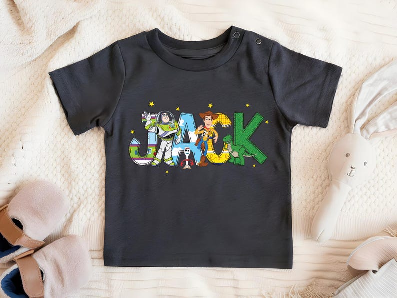 May include: Dark gray short-sleeved t-shirt featuring the name "JACK" in colorful, cartoon-style letters. The letters are decorated with images of characters from the Toy Story movie, including Buzz Lightyear, Woody, and Rex.