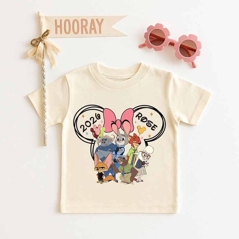 K&ouml;nnte beinhalten: Cremefarbenes T-Shirt mit einem Cartoon-Motiv von Charakteren und dem Text "2020 ROSE" in einem Mickey-Mouse-Ohren-Design. Enth&auml;lt rosa bl&uuml;tenf&ouml;rmige Sonnenbrillen und ein Holzschild mit der Aufschrift "HOORAY".