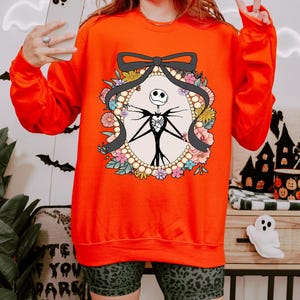 A woman wearing a red sweater with a graphic of a skeleton and floral design, holding a phone and making a peace sign gesture.