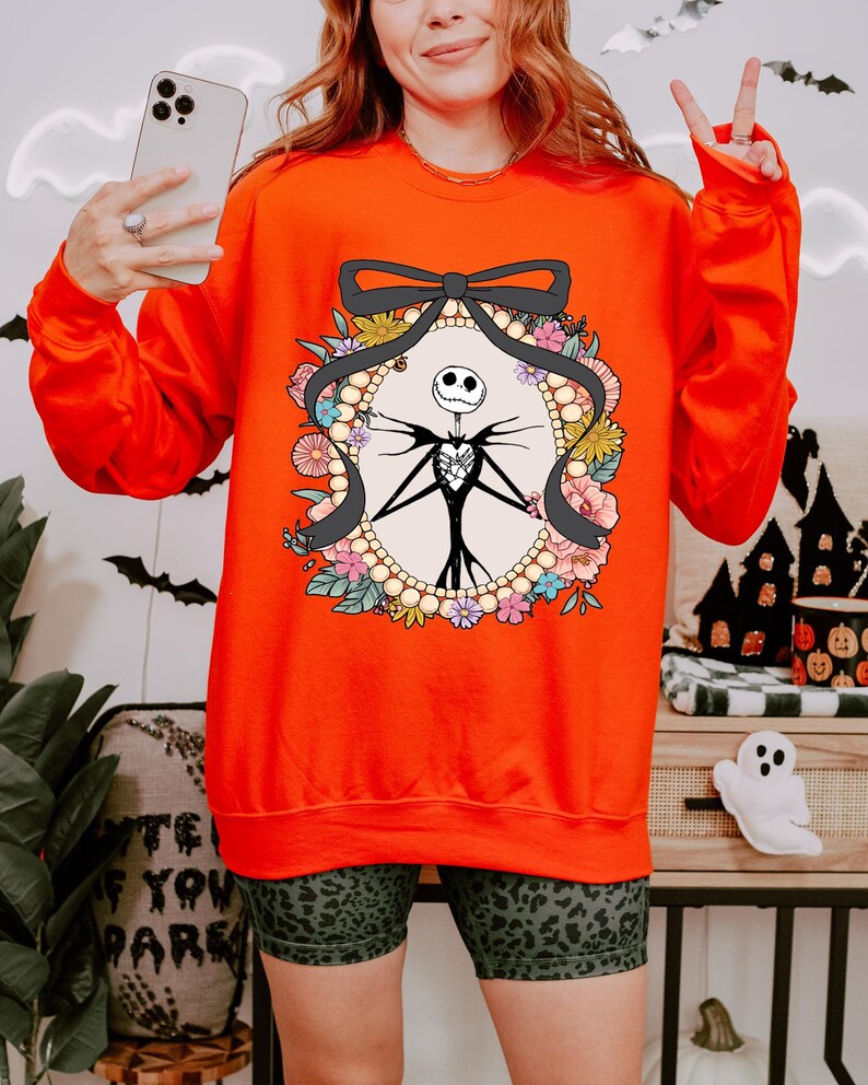 A woman wearing a red sweater with a graphic of a skeleton and floral design, holding a phone and making a peace sign gesture.