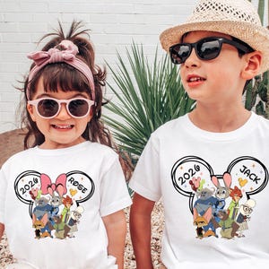 K&ouml;nnte beinhalten: Zwei Kinder tragen wei&szlig;e T-Shirts mit einer Cartoon-Grafik. Die Shirts zeigen ein Design mit dem Text "2026" und Charakterillustrationen. Ein Shirt hat den Namen "Rose", das andere "Jack". Der Junge tr&auml;gt eine Sonnenbrille und einen Strohhut.